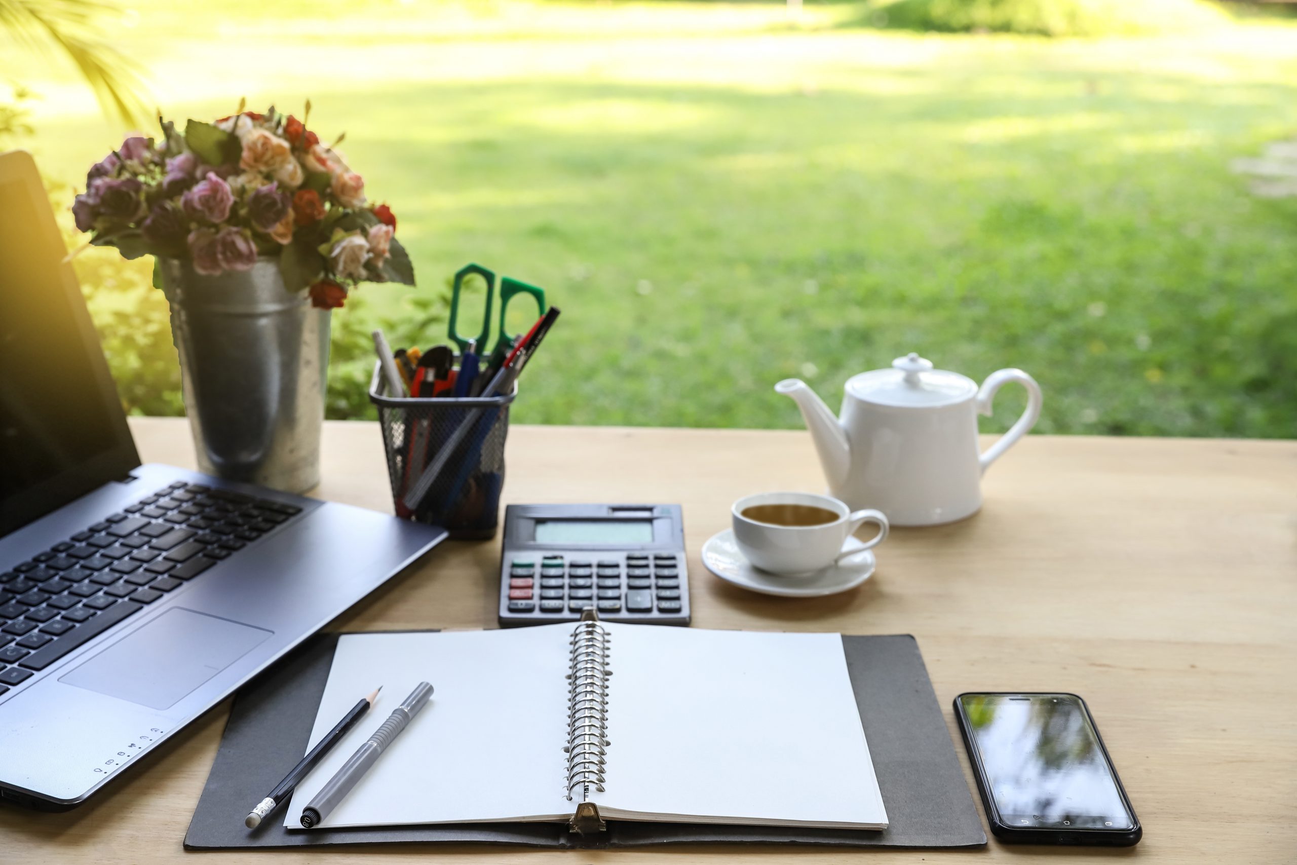 Garden office table with note book ,pen, smartphone,computer business, calculator and cup of tea on green garden background